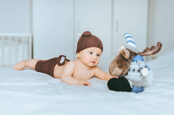 infant child in knitted deer shorts and hat playing with toy moose in bed - Fotó, kép