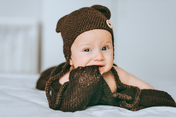 infant child in brown knitted hat biting blanket in bed - Photo, Image