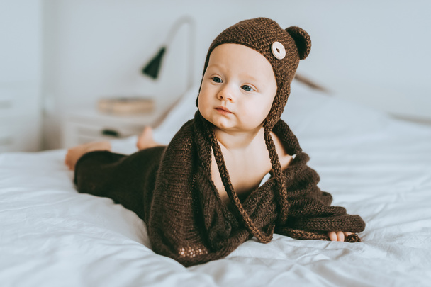 Младенец в коричневой вязаной шляпе и одеяло в постели, смотрит в сторону
 - Фото, изображение