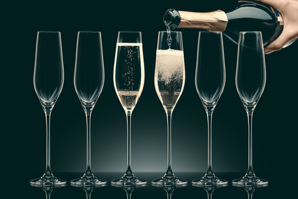 geschnittenes Bild einer Frau, die Champagner aus einer Flasche in sechs transparente Gläser auf Schwarz gießt - Foto, Bild