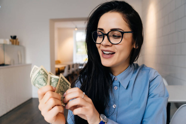 smiling young smart girl with glasses holding dollars in her hands, making a lot of money - Φωτογραφία, εικόνα