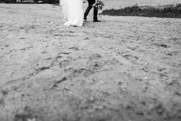 bellissimo matrimonio. gli sposi stanno camminando lungo la sabbia. sposa in un abito di lusso e un bellissimo bouquet. vista posteriore. primo piano di piedi sulla sabbia
 - Foto, immagini