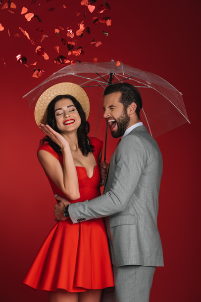 laughing couple hiding under umbrella from falling confetti isolated on red - Foto, Imagen