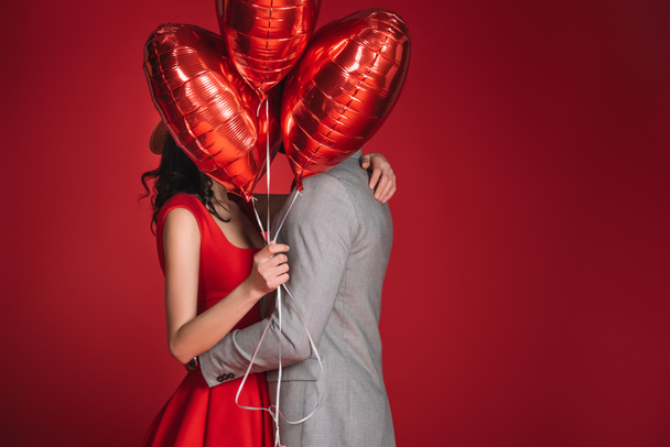 ζευγάρι που καλύπτει πρόσωπα με δέσμη των μπαλονιών που απομονώνονται σε κόκκινο - Φωτογραφία, εικόνα