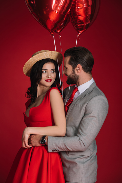 Stylisches Paar mit roten Luftballons, die sich isoliert auf Rot umarmen - Foto, Bild