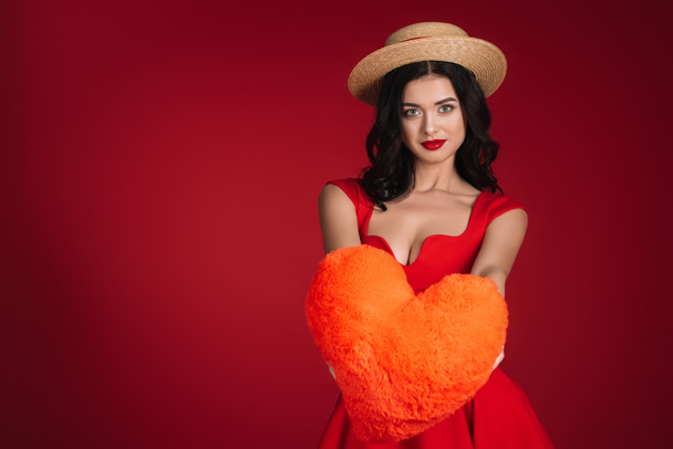 attractive girl in red dress showing heart shaped pillow isolated on red - Fotoğraf, Görsel