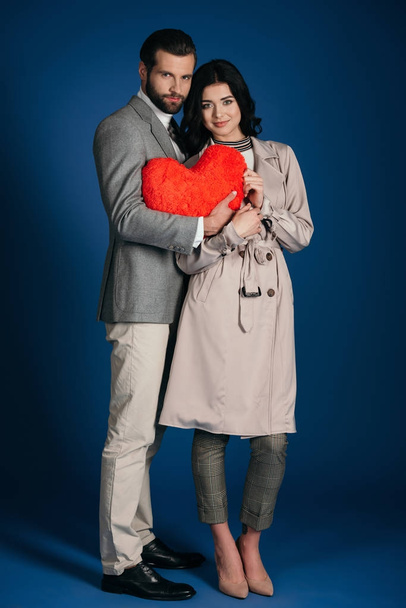 couple holding heart shaped pillow and looking at camera on blue - Fotoğraf, Görsel