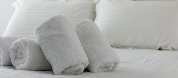 Hotel's bedroom. White fluffy, rolled towels, linen sheets and pillows on a bed. Close up view. - Photo, Image