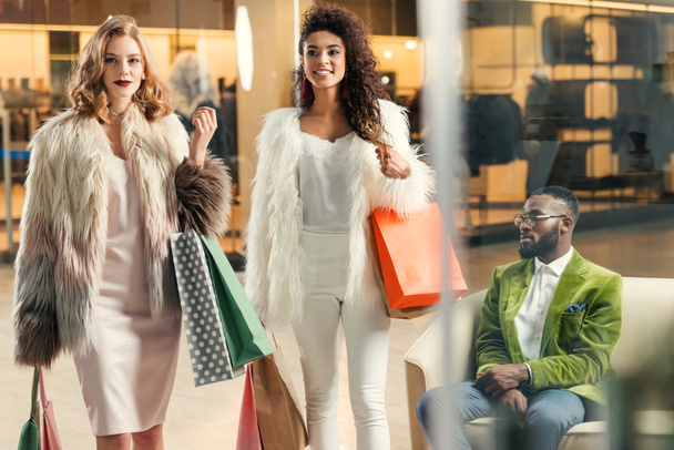 elegáns fiatal többnemzetiségű nők gyalogos bevásárló táskák és afrikai amerikai férfi mögött ülve Mall - Fotó, kép