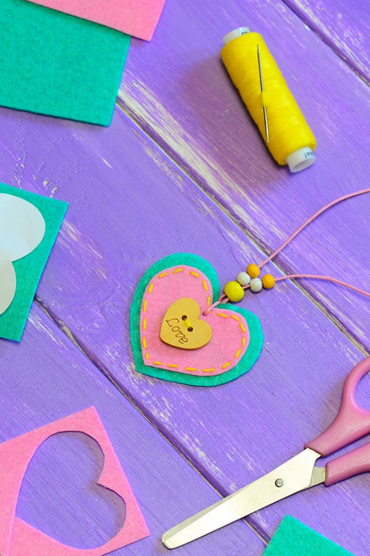 Nice felt heart pendant necklace. Scissors, thread, felt sheets and pieces on a purple wooden background. Valentines Day gift idea. Children creative thinking and activity. Hand sewing projects. Creativity activity for kids. Kids making designs - Photo, Image