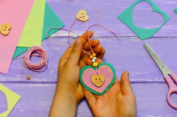 Kind hält eine Halskette mit Herzanhänger in den Händen. Aus Filz, Perlen und Knöpfen bastelte das Kind eine Herzanhängerkette. Nettes Geschenk für Mutti Geburtstag, Muttertag. Valentinstag Geschenk. Handnähprojekte. Kreativitätsaktivität für Kinder - Foto, Bild