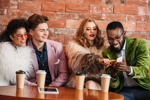 smiling fashionable multiethnic young people using smartphone while drinking coffee - Fotoğraf, Görsel