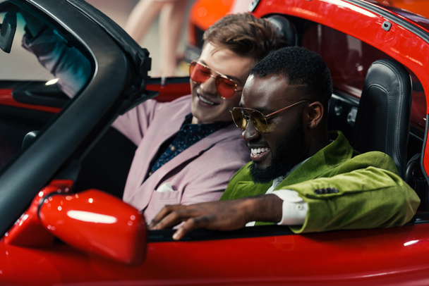 homens multiculturais elegantes felizes sentados em carro novo no showroom
 - Foto, Imagem