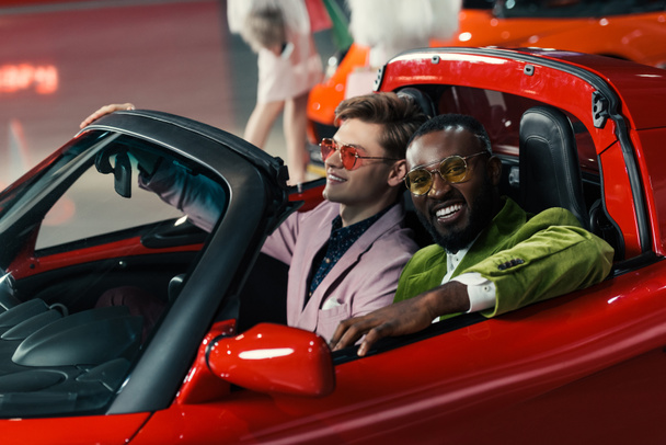 smiling stylish multiethnic men sitting in new car at showroom - Foto, Imagen