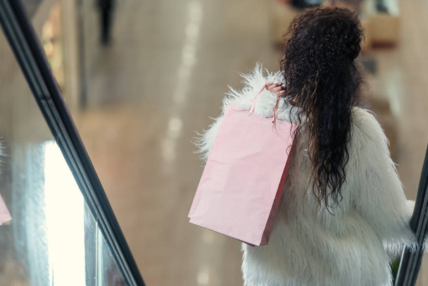 rear view of stylish woman holding shopping bag while riding escalator - Photo, Image