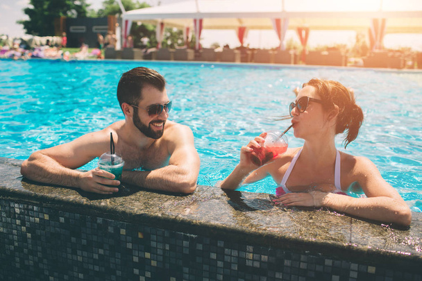 Young couple by the swimming pool. Man and women drinking cocktails in the water. - Photo, Image