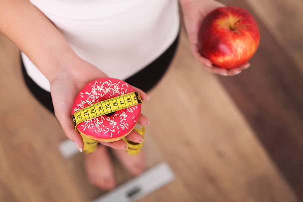 Kevyt. Nainen mittaamalla kehon paino punnitusasteikolla tilalla donitsi ja omena. Makeiset ovat epäterveellistä roskaruokaa. Ruokavalio, Terveellinen syöminen, Elämäntapa. Painonpudotus. Liikalihavuus. Ylhäältä
 - Valokuva, kuva