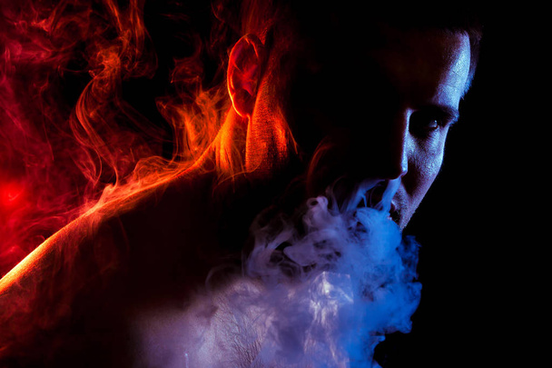 A young man exhales a cloud of colored smoke of red and pink color on a black isolated color all the back is covered with red smoke. Harm to smoking on the example of a young mugger - Foto, afbeelding