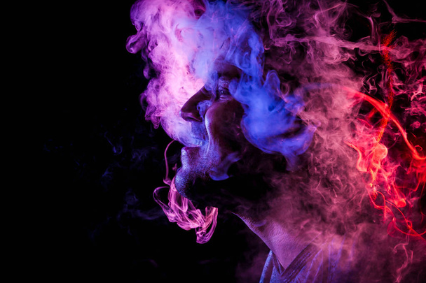 Mann bewundert Dampfschwaden von der elektronischen Zigarette, Wolke von farbigem Rauch von roter und pinkfarbener Farbe auf einem schwarzen isoliert. Schaden durch Rauchen am Beispiel eines jungen Räubers  - Foto, Bild