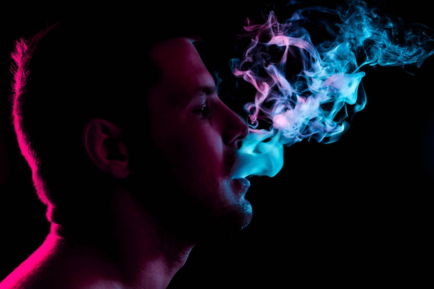 A young man exhales a cloud of colored smoke of red and green color on a black isolated color.  Guy is admiting puffs of steam from the electronic cigarette.  - Valokuva, kuva