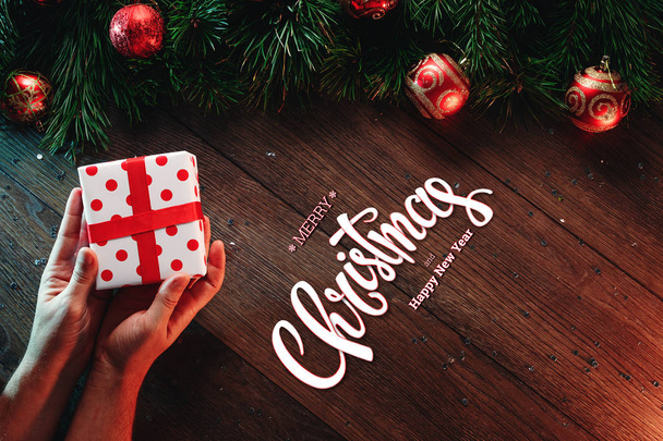 The inscription of Merry Christmas and Happy New Year, male hands hold a gift on a wooden brown table. Christmas card, holiday background. Mixed media. View from above. - Фото, изображение