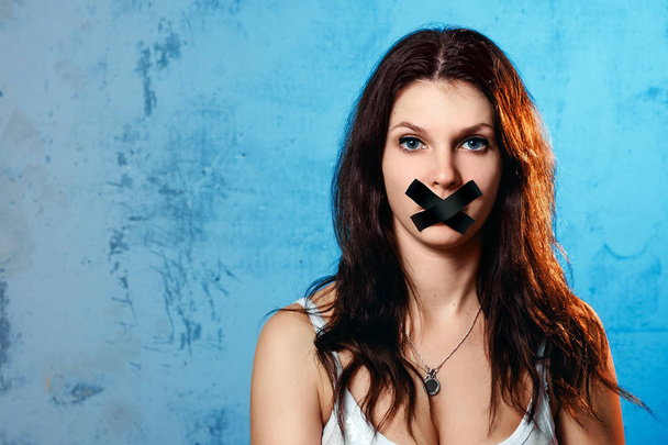 The young woman's mouth is covered with a black ribbon against the blue wall. The concept of domestic violence, domestic mockery of his wife. - Foto, imagen