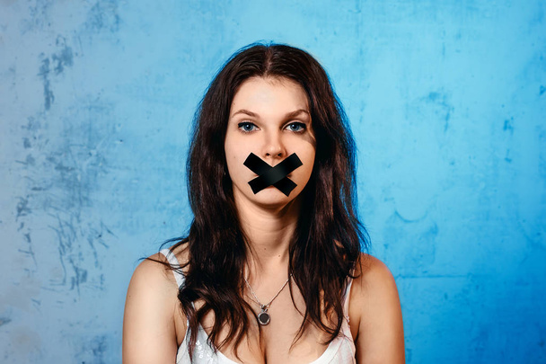 The young woman's mouth is covered with a black ribbon against the blue wall. The concept of domestic violence, domestic mockery of his wife. - Foto, imagen