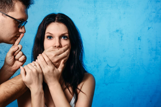 A man closes his mouth against a woman against a blue wall. The concept of domestic violence, domestic mockery of his wife. - Fotografie, Obrázek