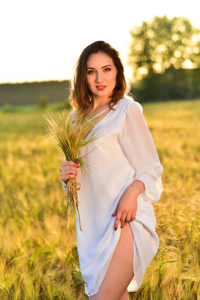 Hermosa joven con vestido blanco en el campo de trigo relajante. Sol resplandeciente
 - Foto, imagen
