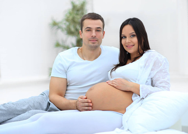 Happy pregnant woman and partner sitting on bed with hands touching tummy - Фото, изображение