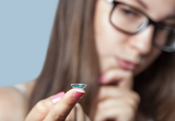A woman in glasses holds a blue contact lens on her finger. Eye care and the choice between the means to improve vision. - Φωτογραφία, εικόνα