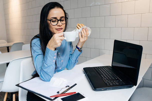 cute young girl in glasses and blue shirt eating a sandwich and coffee in a cafe - Фото, зображення