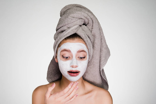 surprised young girl with a towel on her head, on her face a white moisturizing mask - Foto, Imagem