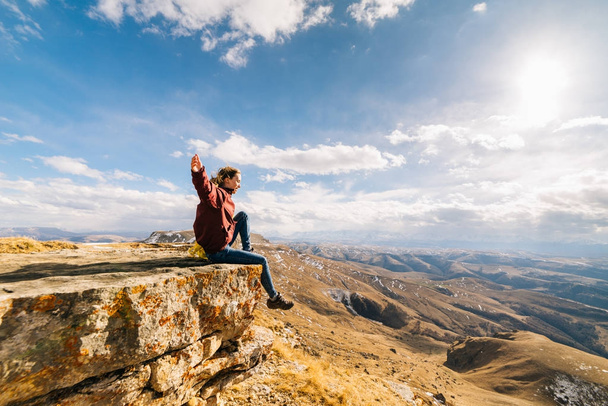 active young girl sits on the edge of the cliff, enjoys the mountain air and the sun - Photo, Image