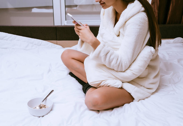 a young girl in a white terry dressing gown and warm socks is sitting in bed and taking pictures of her breakfast - Foto, Bild