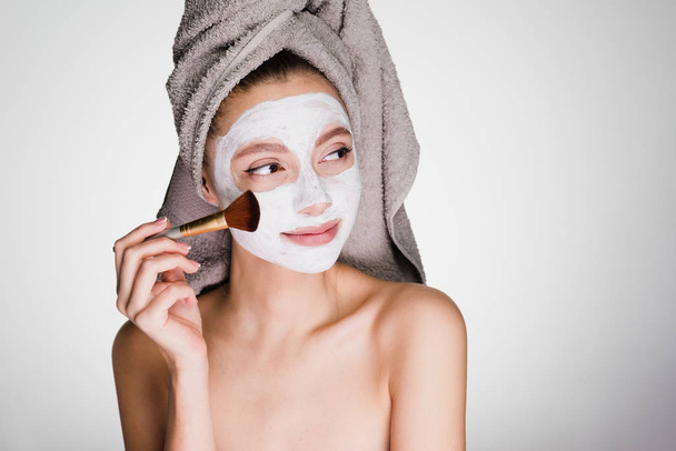 cute attractive girl with a towel on her head applies a nourishing white mask on her face with a brush - Fotografie, Obrázek