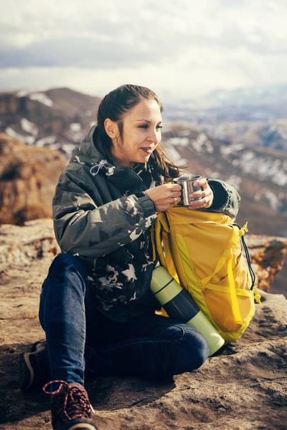 szczęśliwy uśmiechający się dziewczyna siedzi na krawędzi klifu, z żółtym plecak i z przyrodą - Zdjęcie, obraz