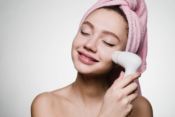 happy young girl with pink towel on her head doing deep cleansing of face with electric brush - Fotó, kép