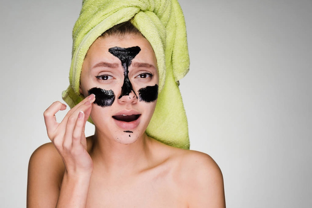 una joven con una toalla verde en la cabeza aplica una máscara de limpieza negra en las áreas problemáticas de su cara
 - Foto, Imagen
