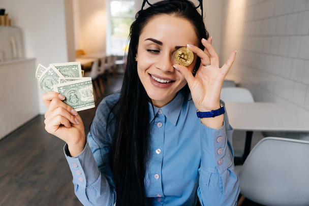 Laughing young girl in blue shirt is holding golden bitcoin and dollars in hands - Фото, изображение