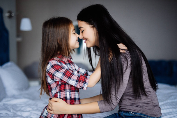 Beautiful woman and her cute little daughter are touching noses and smiling while sitting on bed - Valokuva, kuva