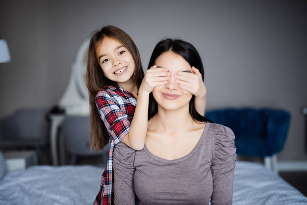 Beautiful young mother and her little daughter are playing on bed at home. Girl is closing her mother eyes - 写真・画像