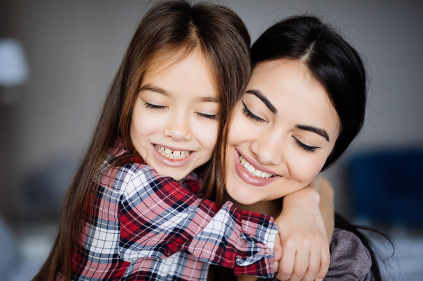Όμορφη νεαρή μητέρα και η κόρη της αγκάλιασμα και χαμογελώντας ενώ κάθεται με κλειστά τα μάτια στο κρεβάτι στο σπίτι - Φωτογραφία, εικόνα