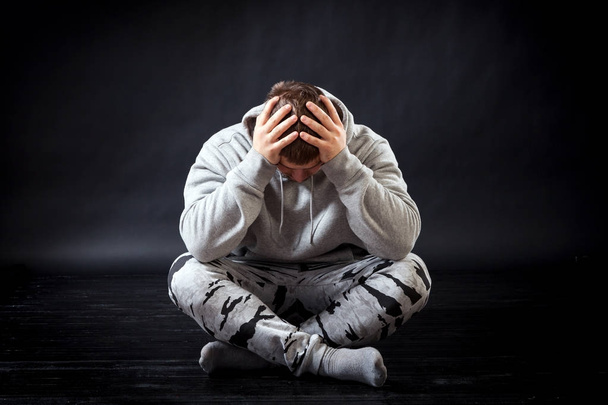 A young sad man in sports clothes sits on the floor and is upset that he is ill, his head hurts, his hands are on his head on a black isolated background. The man is sad with a lot of problems - Foto, immagini