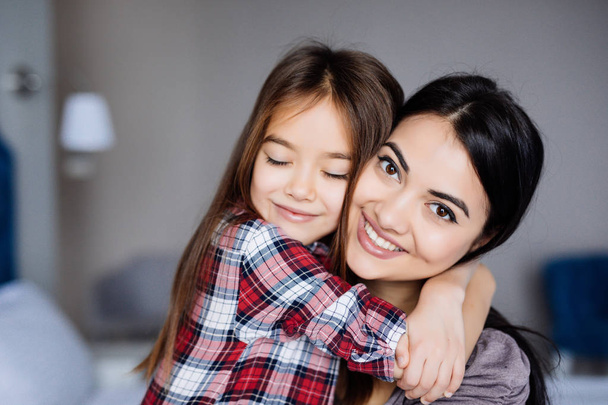 Красивая молодая мать и ее дочь обнимаются и улыбаются, сидя дома на кровати.
 - Фото, изображение