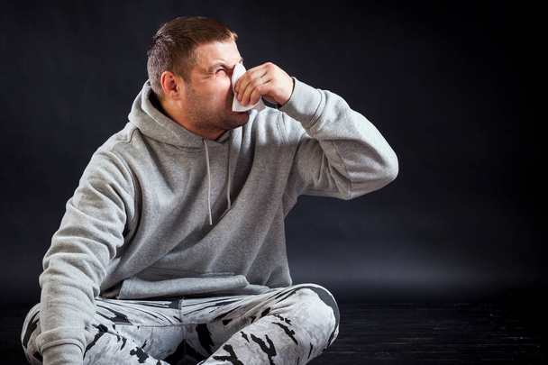 Un joven triste con ropa deportiva está sentado en el suelo enfermo, sosteniendo una servilleta en su mano y sonándose la nariz sobre un fondo negro y aislado. El hombre estornuda
 - Foto, imagen
