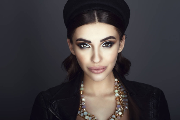 Close up portrait of chic glam dark-haired model with beautiful make up and smooth hair wearing black pillbox hat, leather jacket on naked body and luxurious gem necklace - Photo, Image