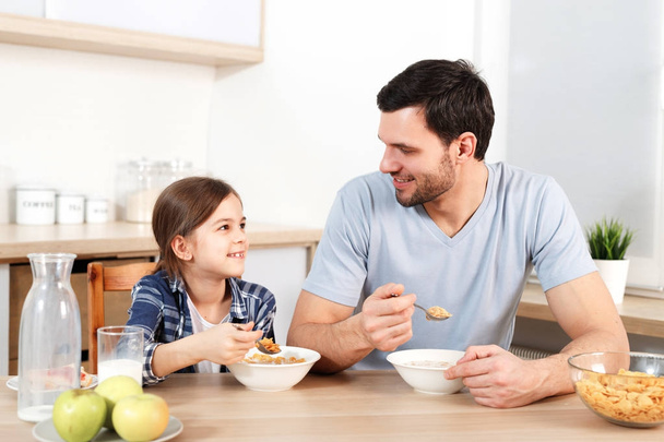 Adorable little kid and her father eat flakes together, have pleasant conversation with each other, sit at kitchen table, eat only healthy food. Parenthood, childhood and eatting concept. - Фото, изображение