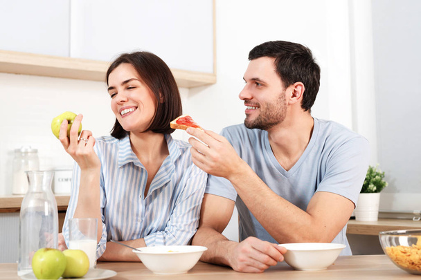 Happy couple spend free time or weekend together at kitchen, glad husband suggests wife to eat snack, she refuses as eats green apple, have breakfast. Family, cooking and relations concept. - Фото, зображення