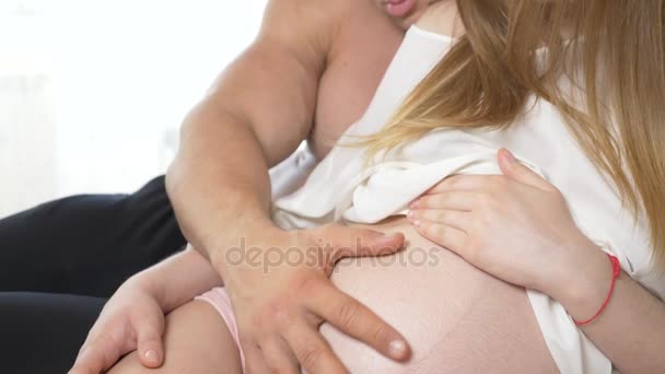 egy gyönyörű fiatal terhes nő és izmos férje feküdni az ágyban, a férj a vasalási feleségének a hasa. 4k, lassú mozgás - Felvétel, videó